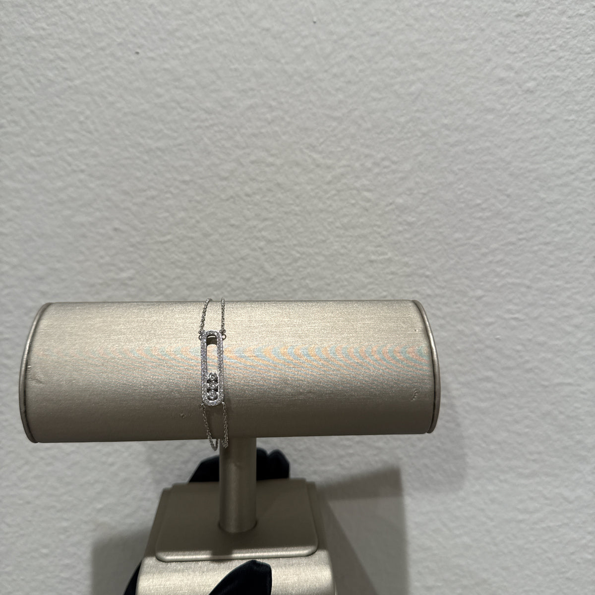 A delicate silver chain bracelet with a small rectangular charm featuring a clear gemstone, displayed on a silver cylindrical jewelry stand against a textured off-white wall.